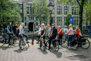 Several people in Amsterdam cycling around the streets. Summer and trees and plants are seen outside the buildings.
