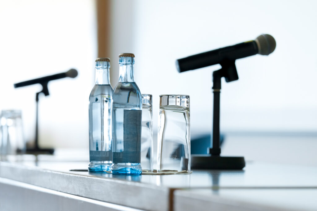 Lectern with water bottles, glasses and microphone. Focus on glasses. Some grain visible.